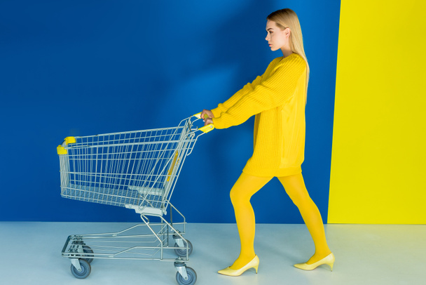 Elegant blonde woman pushing shopping cart on blue and yellow background - Foto, afbeelding
