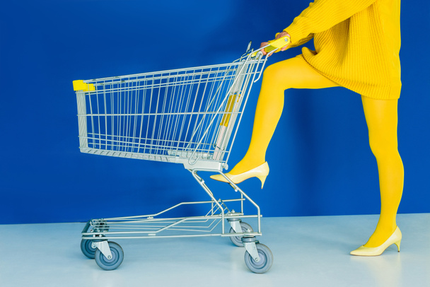 Cropped view of woman leaning on shopping cart on blue background - Foto, Imagem