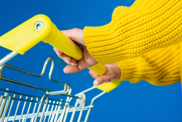 Cropped view of female hands on shopping cart handle isolated on blue background - Fotografie, Obrázek
