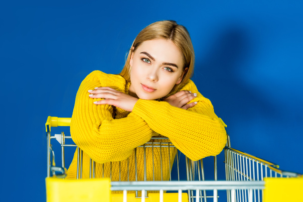 Elegant blonde woman in yellow garment leaning on shopping cart on blue background - Photo, Image