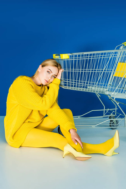 Attractive young girl sitting on floor and leaning on shopping cart on blue background - Φωτογραφία, εικόνα