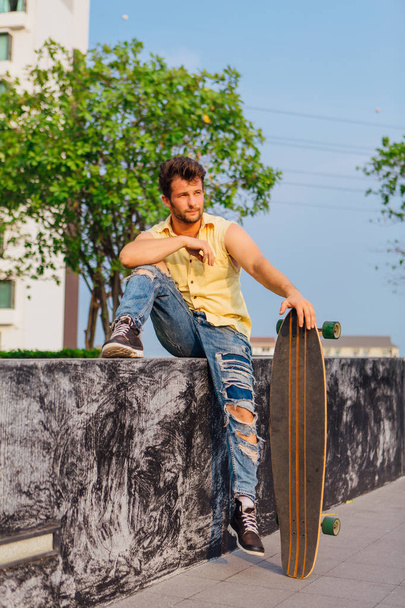Young handsome man with beard sitting with longboard on the street. - Zdjęcie, obraz