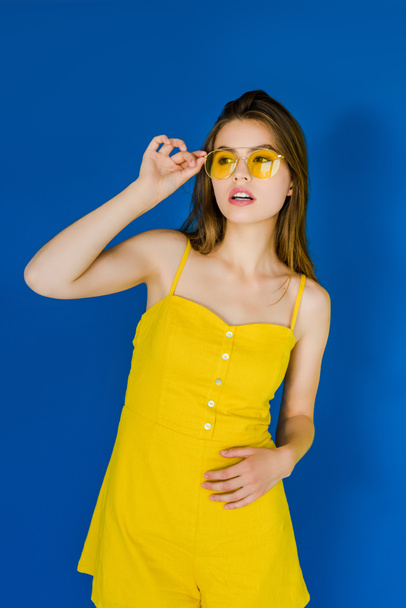 Attractive young girl wearing yellow sunglasses posing on blue background - Foto, Imagen
