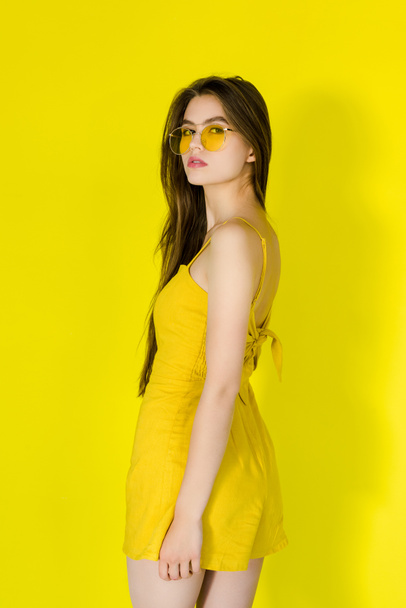 Female fashion model in yellow dress and sunglasses posing on yellow background - Foto, Imagen