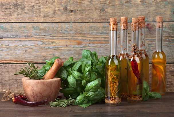 Olijfolie met verschillende kruiden en kruiden op een oude houten tafel. Flessen van olijfolie met basilicum en houten mortel. - Foto, afbeelding