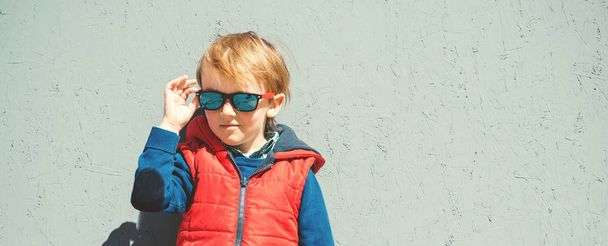 Stylish kid in trendy sunglasses. Kids fashion. Cute little blondy boy in red jacket standing over the grey wall outdoors in sunny day. Autumn, spring kids fashion. Kids sunglasses. Hipster style. - Foto, Imagem
