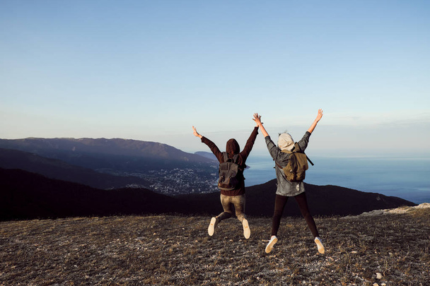 Two girls traveler stand on the top of the mountain, look down, jump, rejoice and dance. - Foto, afbeelding