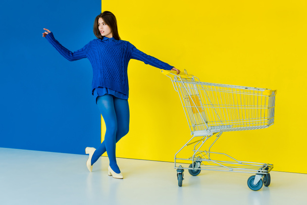 elegante brünette Frau in blauem Pullover posiert am Einkaufswagen auf blauem und gelbem Hintergrund - Foto, Bild