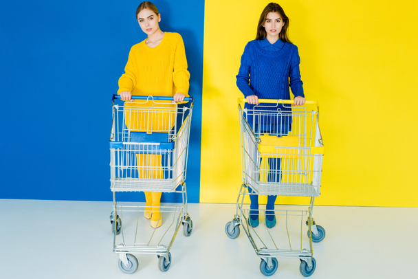 Attractive young girls in fashion clothes pushing shopping carts on blue and yellow background - Photo, Image