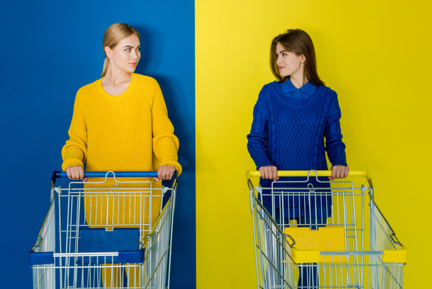 Mosolygó fiatal nő a bevásárlókocsik néztek egymásra, a kék és a sárga háttér - Fotó, kép