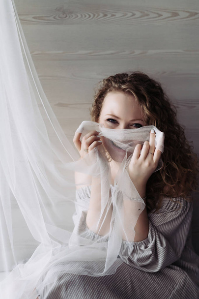 a funny curly-haired woman in a dress sits at the window, covered her face with a curtain, laughs - Foto, afbeelding