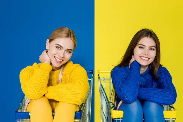 Attractive young girls having fun sitting in shopping carts isolated on blue and yellow background - Photo, Image