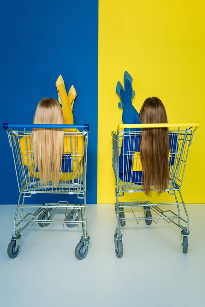 Achteraanzicht van vrouwelijke fashion modellen zitten in winkelwagentjes op blauwe en gele achtergrond - Foto, afbeelding