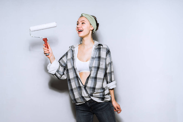 joyful young girl in plaid shirt colors the wall in white with a roller, smiling - Foto, imagen