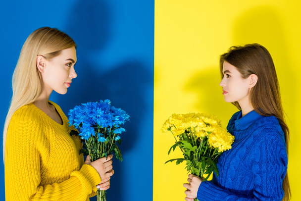 Models halten Chrysanthemensträuße in der Hand und betrachten einander auf blauem und gelbem Hintergrund - Foto, Bild