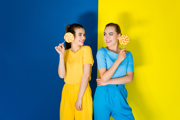 Beautiful blonde and brunette girls holding lollipops on blue and yellow background - Fotó, kép