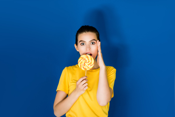 Beautiful surprised brunette girl holding lollipop on blue background - Foto, Imagen