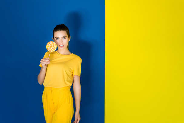 Attractive young girl in yellow outfit holding lollipop on blue and yellow background - Foto, immagini