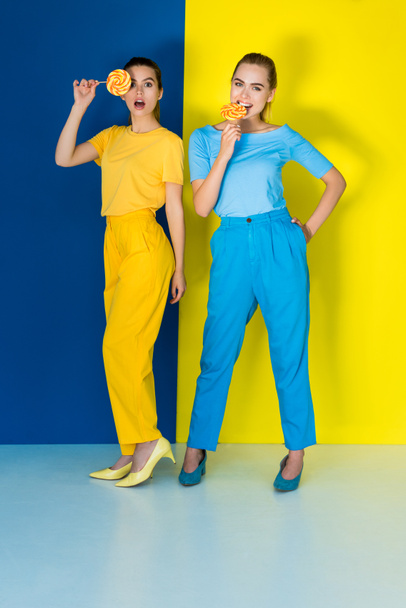 Eleganti donne alla moda mangiare lecca-lecca su sfondo blu e giallo
 - Foto, immagini