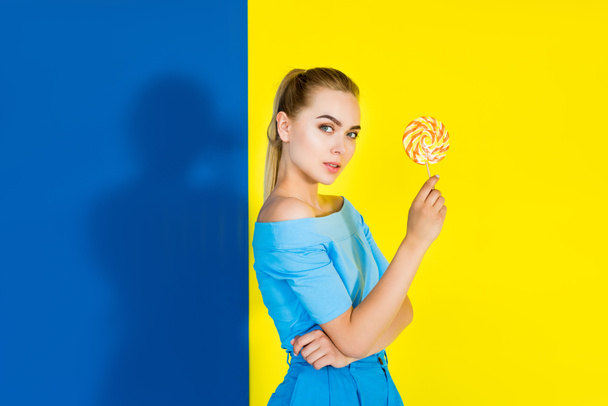 Atraktivní mladá dívka držící lízátko a při pohledu na fotoaparát na modré a žluté pozadí - Fotografie, Obrázek