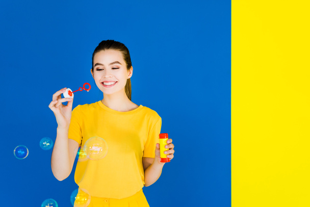 Krásná brunetka vyfukuje bubliny izolované na modré a žluté pozadí - Fotografie, Obrázek
