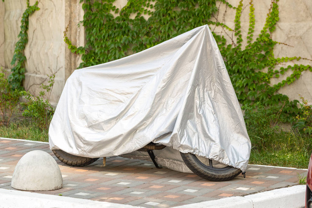 Motocicleta cubierta con chaqueta protectora gris. Scooter con escudo de tela aparcado en el pavimento cerca de la pared de hormigón. Estacionamiento impermeable al aire libre durante el viaje
 - Foto, imagen