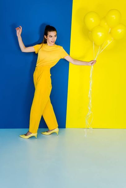Beautiful brunette girl holding bunch of yellow balloons on blue and yellow background - Фото, изображение