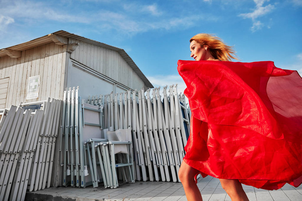 Retrato ao ar livre dinâmico de mulher loira atraente em vestido voador vermelho, andando perto de edifício de madeira branca na costa
   - Foto, Imagem