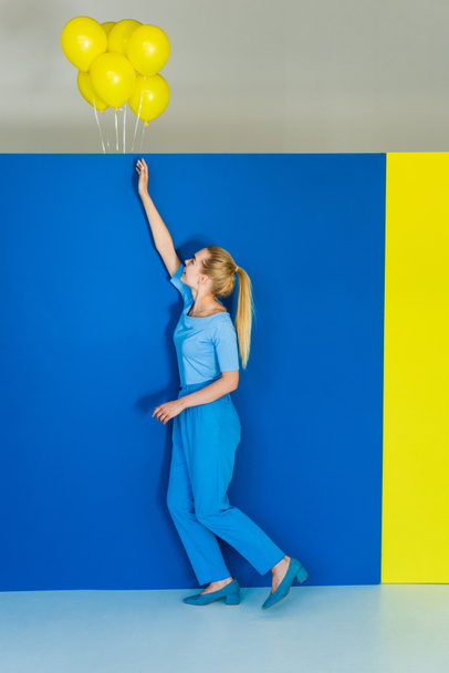 schöne blonde Mädchen greift nach gelben Luftballons auf blauem und gelbem Hintergrund - Foto, Bild