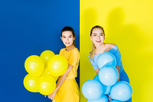 Mulheres elegantes e elegantes sorrindo e segurando balões de festa em fundo azul e amarelo
 - Foto, Imagem
