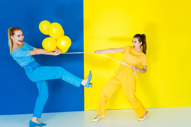 Blondýna a bruneta dívky bojují za balónky na modré a žluté pozadí - Fotografie, Obrázek