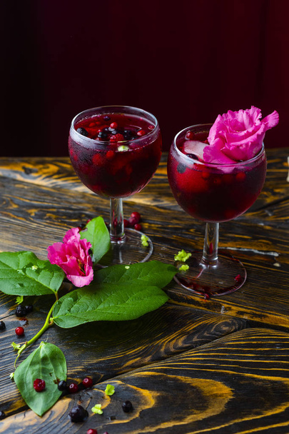 Two glasses full of red drink with berries, ice and a pink flowers, beside which lie berries and a branch of green tropical leaves on a vintage wooden table. Copy space. Commercial design. - Zdjęcie, obraz