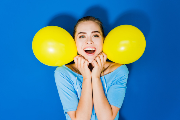 Muchacha joven atractiva sonriendo y sosteniendo globos por su cara sobre fondo azul
 - Foto, imagen