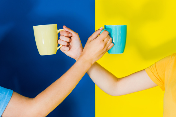 Cropped view of colorful cups in women hands on blue and yellow background - Photo, Image