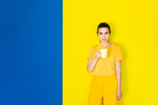 Beautiful brunette girl holding yellow cup on blue and yellow background - Photo, Image