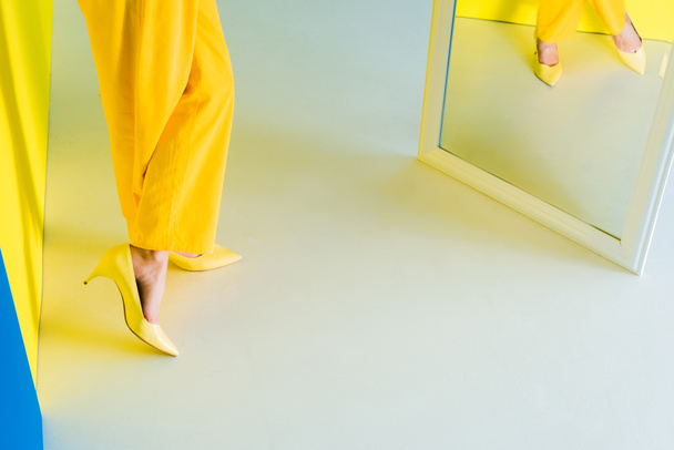 Vista recortada de la mujer en traje amarillo de pie delante del espejo sobre fondo azul y amarillo
 - Foto, Imagen