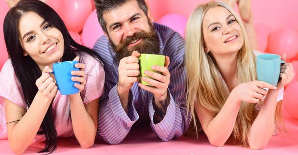 Man and women in domestic clothes, pajamas. Threesome relax in morning with coffee. Lovers drinking coffee in bed. Lovers in bed concept. Man and women, friends on happy faces lay, pink background - Valokuva, kuva