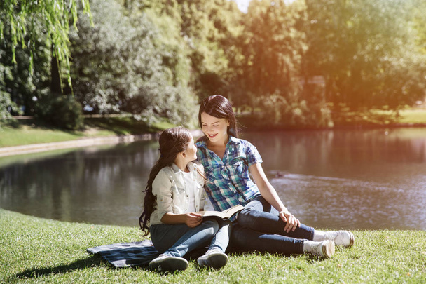 Pretty girls are sitting at the edge of small lake in park. They are looking at each other. Kid is holding opened book on her lap. - Zdjęcie, obraz