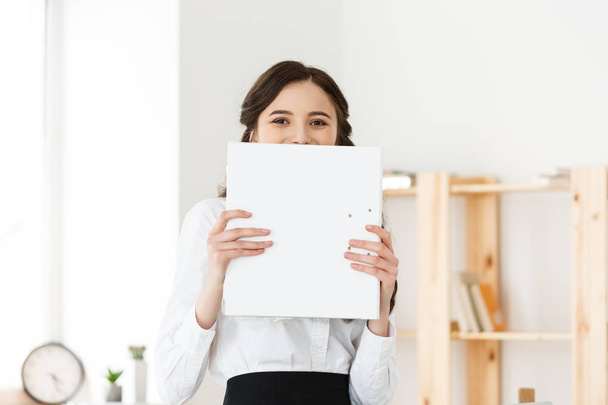 Young woman with surprised or happy eyes peeking out from behind paper poster. Businesswoman holding big white banner in modern office. - Photo, Image