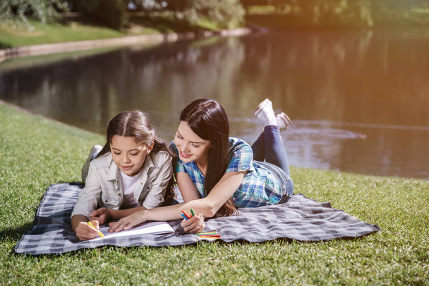 Happy mom is lying on blanket with her daughter. She holds her hand on piece of paper. Girl is drawing with yellow marker. They are smiling and looking down. - Foto, Bild
