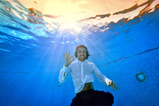 Usměvavý mladík s mikrofonem na hlavě plave a představuje pod vodou v bazénu v bílé košili proti žluté slunce. Dívá se na kameru. Portrét. Podvodní fotografie. - Fotografie, Obrázek