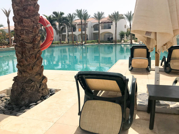 Relax-Pool mit klarem, blauem Wasser mit Liegestühlen zum Sonnenbaden mit Sonnenschirmen vor dem Hintergrund eines Palmenstammes mit Rettungsring in einem meertropischen Resort im All-Inclusive-Hotel. - Foto, Bild