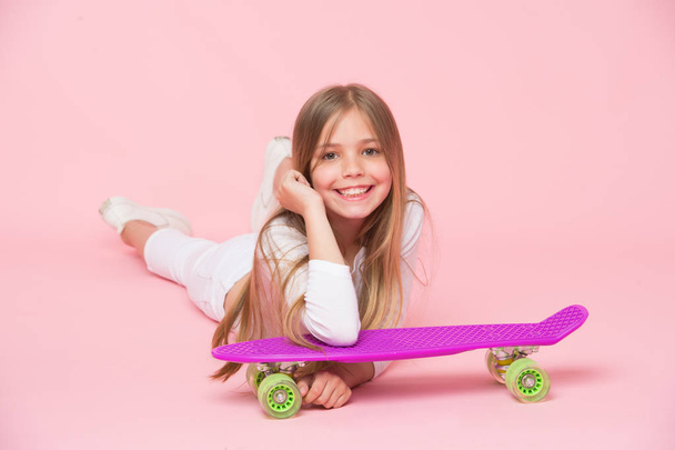 Kid in white outfit lying on floor. Girl with long blond hair isolated on pink background. Child with cute smile leaning on violet skateboard. Teenager having fun, relaxation concept - Photo, image