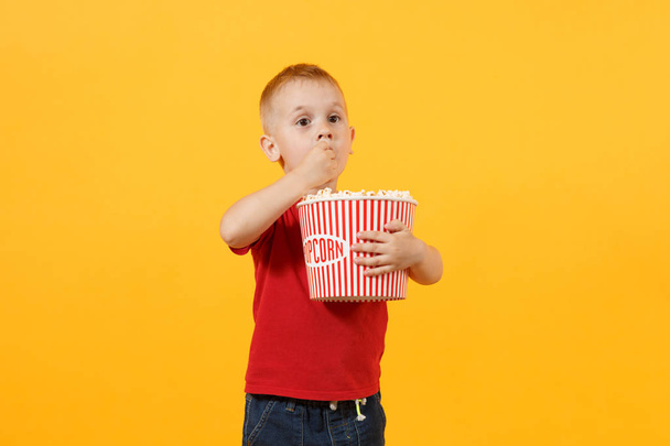 Маленький милий веселий хлопчик 3-4 роки в червоній футболці тримає відро для попкорну, їсть фаст-фуди ізольовані на жовтому тлі. Дитяча концепція дитячого способу життя. Нездоровий фастфуд. Копіювати простір
 - Фото, зображення