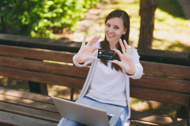 Young successful businesswoman holding credit card. Woman sitting on bench working on modern laptop pc computer in city park in street outdoors on nature. Mobile Office. Freelance, business concept - Foto, imagen