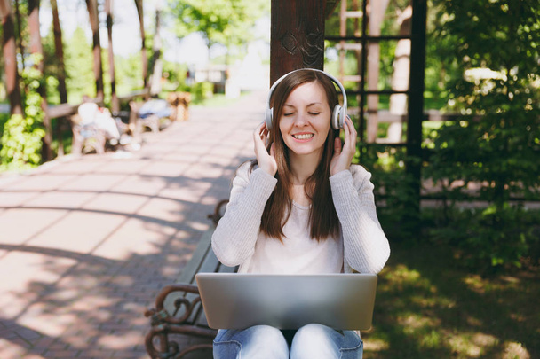 Молодая успешная бизнесвумен в легкой повседневной одежде. Женщина, работающая на современном ноутбуке, слушает музыку в наушниках на голове на улице. Мобильный офис. Концепция внештатного бизнеса
 - Фото, изображение