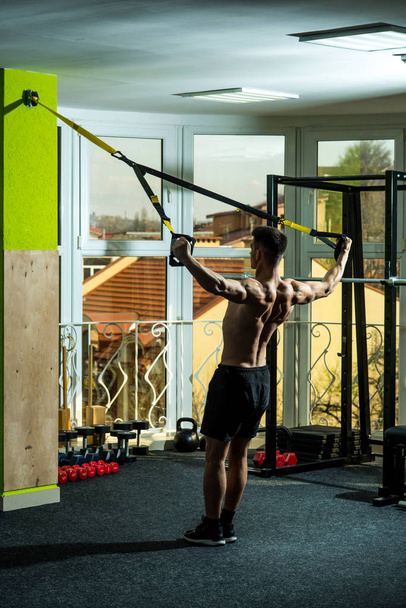 Man with nude torso, and muscular back in gym enjoy training, trx. Sport and gym concept. Man with torso, sportsman, athlete, muscular macho does exercise with trx loops, window on background - Foto, Bild