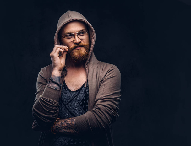 Nachdenklicher Rotschopf-Hipster mit Vollbart und Brille in Kapuzenpulli und T-Shirt posiert mit Hand am Kinn in einem Studio. isoliert auf dunklem Hintergrund - Foto, Bild