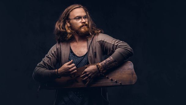 Redhead hipster male with long luxuriant hair and full beard dressed in casual clothes playing on a Russian traditional musical instrument - gusli. Isolated on the dark background.  - Photo, Image