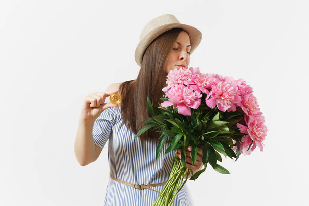 Young woman in dress, hat holding bitcoin, coin of golden color, bouquet of beautiful pink peonies flowers isolated on white background. Business, delivery, online shopping, virtual currency concept - Photo, Image